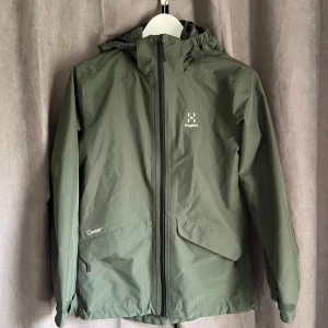 Grön vindjacka från Haglöfs - Säljer en grön vindjacka från Haglöfs med huva och dragkedja framtill. Jackan har stora fickor med dragkedja och meshfoder på insidan. Perfekt för blåsiga dagar och outdoor-äventyr. Liten skada brevid dragkedjan, se andra bilden, men mycket bra skick i övrigt. Originalpris 1500kr.