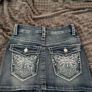 Jeanskjol med broderade fickor - Snygg kort jeanskjol i blå denim med broderade fjärilar och pärldetaljer på bakfickorna. Klassisk femficksmodell med knapp och dragkedja framtill. Perfekt för dig som gillar unika detaljer och cool stil.