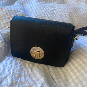 Svart axelväska i skinn med gulddetalj - Snygg svart axelväska i skinn med en rund guldfärgad låsdetalj framtill. Väskan har en justerbar axelrem och stilren, boxig form. Perfekt storlek för det viktigaste och ger en clean look till outfiten. Liten skrapmärke på knäppet men det syns inte när väskan är stängd.