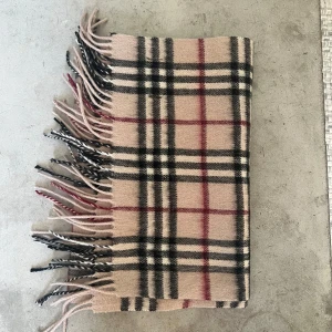 Beige rutig halsduk från Burberry - Säljer en klassisk beige halsduk från Burberry i 100% kashmir. Den har det ikoniska rutiga mönstret i svart, vitt och rött samt snygga fransar i ändarna. Perfekt accessoar för att lyfta din outfit med lyxig känsla och tidlös stil.