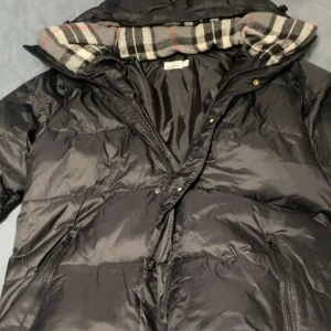 Svart dunjacka från Burberry - Säljer en svart dunjacka från Burberry med klassiskt rutigt foder i huvan. Jackan har dragkedja, knappar och två fickor framtill. Stor Burberry-patch på ärmen och quiltad design. Perfekt för kalla dagar och riktigt snygg streetstil.Säljer nu min oanvända Burberry vinterjacka då den inte var min smak. Jackan är stilren och går med de flesta outfits. För fler frågor skriv privat. Priset kan diskuteras 