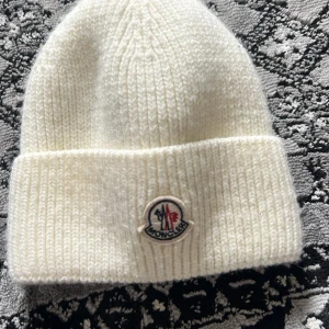 Vit stickad mössa från Moncler - tyvärr måste jag sälja denna efterlängtade varan eftersom att den är för liten på mig, jag insåg det när jag beställde den. Säljer en stilren vit stickad mössa från Moncler med klassisk uppvikt kant och broderad logotyp framtill. Mössan är ribbstickad och tillverkad i mjukt material som håller dig varm under kyliga dagar. Perfekt accessoar för vintern!