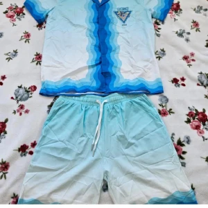 Casablanca blått set med vågmönster - Säljer ett fräscht set från Casablanca Tennis Club med skjorta och shorts. Setet har ett vågigt blått och vitt mönster, tryckt logga på både skjorta och shorts samt bekväm resår och snörning i midjan. Perfekt för sommardagar och chill häng.