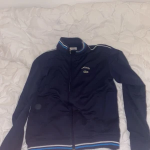 Marinblå Lacoste ziptröja med ränder - Snygg marinblå ziptröja från Lacoste med broderad logga och krokodil på bröstet. Tröjan har hel dragkedja, ståkrage och vita samt blå ränder på krage och muddar. Perfekt för en sportig och stilren look.