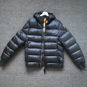 Svart dunjacka Parajumpers Sheen L - Säljer en svart dunjacka från Parajumpers, modell Sheen. Självklart är jackan äckta, medföljer en galge och jackan är köpt i vintras.  Jackan har en glansig finish, huva och dragkedja framtill. Klassisk patch på ärmen och ribbade muddar. Perfekt för kalla dagar när du vill hålla stilen.