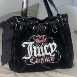 Juicy Couture svart axelväska i sammet - Juicy Couture axelväska i svart sammet med glittriga broderier och rosa krona framtill. Väskan har svarta handtag i skinnimitation, snörning upptill och innerfoder med tryckt logga. Snygg detalj med rosett på sidan och gott om plats för allt du behöver. Köpt på second hand så kan ej lova att den är äkta men alla test tyder på det. Den rosa kronan är lite dåligt skicka (se bild 5). Det är bara att kontakta om du har fler frågor eller funderingar💕