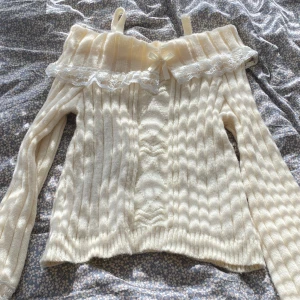 Offshoulder ribbad stickad tröja med spets - Supersöt offshoulder stickad tröja i ljusbeige med ribbat mönster, spetskant och liten rosett framtill. Långa ärmar och axelband för extra detalj. Perfekt för dig som gillar romantisk och feminin stil.