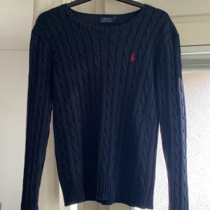 Mörkblå kabelstickad tröja Polo Ralph Lauren - Kabelstickad mörkblå tröja från Polo Ralph Lauren med klassisk röd logga broderad på bröstet. Tröjan har rund halsringning, långa ärmar och ribbade muddar. Perfekt för dig som gillar en tidlös och stilren look.