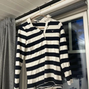 Randig pikétröja från GANT XS - Snygg långärmad pikétröja från GANT i svart och vitt. Tröjan har breda ränder, klassisk krage och knappar vid halsen. Tillverkad i mjuk bomull, perfekt för en chill och stilren look.