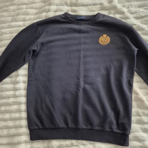 Svart sweatshirt från GANT med emblem - Svart sweatshirt från GANT med guldfärgat broderat emblem på bröstet. Klassisk rund halsringning och ribbade muddar vid ärmslut och nederkant. Perfekt för en stilren och avslappnad look.