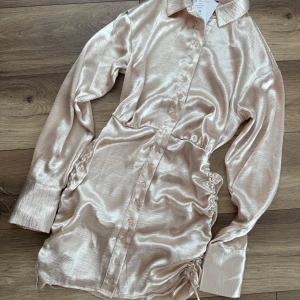 Champagnefärgad skjortklänning i satin - Superfin kort skjortklänning i glansig champagnefärgad satin från Gina. Klänningen har långa ärmar, knappar hela vägen framtill, krage och snygga snörningsdetaljer i sidorna som ger en cool rynkad effekt. 