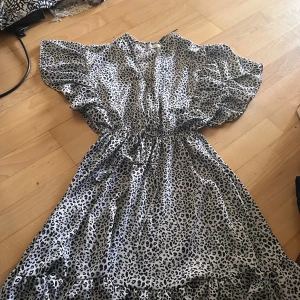 Leopardmönstrad omlottklänning - Snygg leopardmönstrad klänning med volangdetaljer och omlottmodell. Klänningen har korta, vida ärmar och markerad midja med knytband. Perfekt för dig som vill sticka ut med ett trendigt djurmönster.