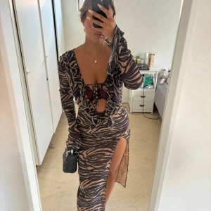 Zebramönstrad långklänning med slits - Säljer en långärmad zebramönstrad långklänning i beige och svart med djup v-ringning, volangdetaljer och snörning framtill. Klänningen har en hög slits på ena sidan och är figurnära med transparent känsla. Perfekt för dig som vill sticka ut!