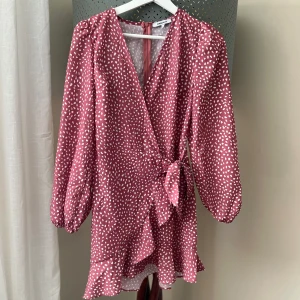 Rosa/röd omlottklänning från Chiquelle - Söt rosa/röd omlottklänning från Chiquelle med vita prickar. Klänningen har långa ärmar och en knytning i midjan.