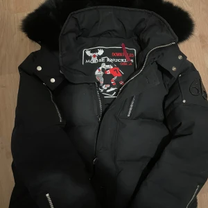 Svart dunjacka från Moose Knuckles - Hallå, dags för mig att sälja mina vinter kläder och därmed min fina moose knuckles. Har använt den under vintern ett par gånger men använde mig av andra jackor då den var lite för varm ibland. Ny kostar den ung 12000kr.   Loggan är lite sliten men går att fixa och som du ser på bild 3 ovanför märket så ser man att bandet är löst från ena sidan. Utöver detta har den inga problem.  Kan tänka mig att gå ner i pris vid snabb affär.                   Kom gärna med egna bud! Kvitto finns!
