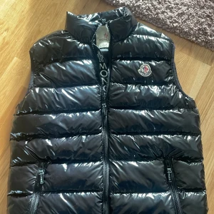 Svart dunväst från Moncler snälla köp - Snygg svart dunväst från Moncler med glansig yta och dragkedja framtill. Västen har en broderad logotyp på bröstet och praktiska fickor med dragkedja. Perfekt för kyligare dagar. Den är bra skik och snälla köp jag vill verkligen sälja kan byta något annars om man vill ?