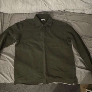 Olivgrön pikétröja - Säljer en stilren olivgrön pikétröja med långa ärmar. Perfekt för en avslappnad men ändå snygg look. Tröjan har en klassisk krage och knappar vid halsen. Storlek s