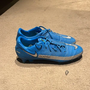 Blå Nike Phantom fotbollsskor - Snygga blå Nike Phantom fotbollsskor med vit swoosh och snörning. Skorna har en mönstrad ovandel och gröna dobbar. Säljer dessa nu då de är lite för små för att spela med under längre tid. Använda cirka 2 månader och i väldigt gott skick.