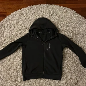 Svart hoodie från Sail Racing - Säljer en svart hoodie från Sail Racing med dragkedja och huva. Tillverkad i en blandning av bomull och polyester, vilket gör den både bekväm och hållbar. Perfekt för kyliga dagar. Märkeslogga på bröstet ger en sportig touch. Nästan aldrig använt. 