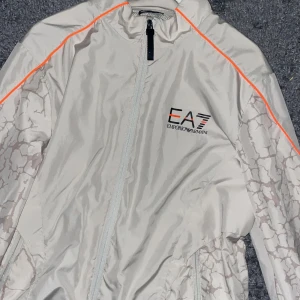 Vit vindjacka från Emporio Armani - Säljer en stilren vit vindjacka från Emporio Armani med ett unikt mönster och orange detaljer. Jackan har dragkedja framtill och EA7-logga på bröstet. Perfekt för en sportig look!