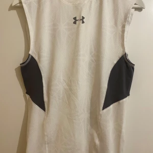 Vit ärmlös kompressionstopp från Under Armour - Säljer en vit ärmlös kompressionstopp från Under Armour med gråa sidopaneler. Linnet är I strlk M