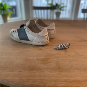 Valentino Garavani sneakers, orginal snören som följs med - Snygga Valentino Garavani sneakers i vitt och grått. Skorna har en bred grå rem över ovansidan och är tillverkade i skinn. Perfekta för en stilren look. 