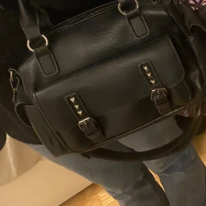 Svart axelväska med nitar - Snygg svart axelväska med coola nitar på framsidan. Väskan har både handtag och en justerbar axelrem för flexibel användning. Axelremmen kan man ta av. Den har en praktisk dragkedja upptill och flera fack inuti för enkel organisering.