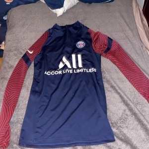 Blå PSG fotbollströja från Nike - Snygg blå PSG fotbollströja från Nike med röda mönstrade ärmar. Tröjan har en dragkedja vid halsen och PSG-loggan på bröstet. Tillverkad i Dri-FIT-material för optimal komfort. Perfekt för fotbollsfans som vill visa sitt stöd för laget.