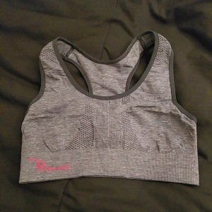 Grå sport bh från Panos - Snygg grå sport bh från Panos med rosa logga. Toppen har en tight passform och är tillverkad i ett bekvämt syntetmaterial. Perfekt för träning med sitt andningsbara tyg och stilrena design.