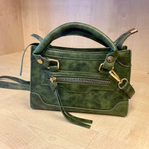 Grön handväska - Snygg grön axelväska som man kan ta av bandet på så det blir en handväska. Väskan har guldfärgade detaljer och en ficka i mitten och en liten ficka framtill💕