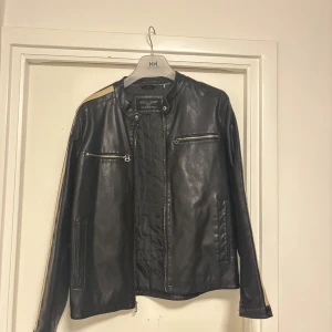 Svart skinnjacka från Wilsons Cycle - Snygg svart skinnjacka från Wilsons Cycle med dragkedja och coola detaljer. Jackan har långa ärmar med ränder i beige och orange längs sidorna. Perfekt för en stilren look. Äkta läder!!