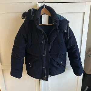 Mörkblå dunjacka från Polo Ralph Lauren - Säljer en snygg mörkblå dunjacka från Polo Ralph Lauren. Jackan har en quiltad design med dragkedja och knappar framtill. Insidan är fodrad med ett stilrent rutigt mönster. Perfekt för kalla dagar med sin varma och bekväma passform.