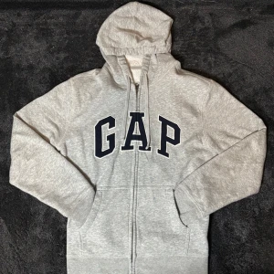 Grå hoodie från GAP - Säljer en klassisk grå hoodie från GAP med dragkedja och stor logga på bröstet. Tröjan har en justerbar huva och långa ärmar. Perfekt för en avslappnad stil. Tröjan är storlek M.