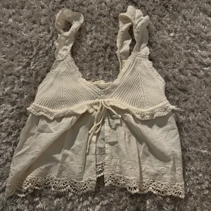 Creme färgad stickad topp från Zara - Charmig vit stickad topp från Zara med spetsdetaljer och knytning framtill. Toppen har en vacker volangkant och är perfekt för varmare dagar. Den är tillverkad i ett mjukt material som ger en romantisk känsla.