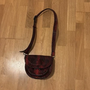 Röd axelväska med ormmönster NA-KD - Snygg röd axelväska med ormmönster och justerbar axelrem. Väskan har en rundad form och ett lock med knäppning. Perfekt för att ge en edgy touch till din outfit.