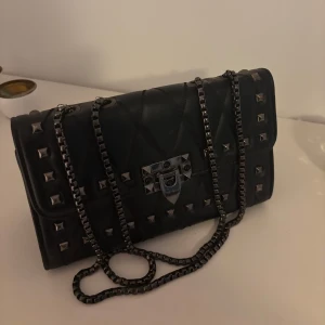 Svart axelväska med nitar - Snygg svart axelväska med quiltad design och coola nitar. Väskan har en kedjerem och ett metallspänne framtill. Perfekt för att ge din outfit en edgy touch.
