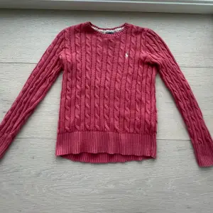 Säljer en snygg rosa stickad tröja från Ralph Lauren med kabelstickat mönster. Tröjan har långa ärmar och en rund halsringning. Perfekt för kyligare dagar och ger en klassisk look. 🌸