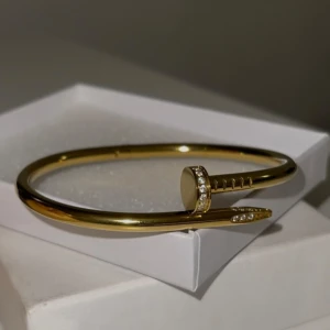 Guldarmband med diamantdetaljer - Elegant guldarmband designat som en böjd spik med gnistrande diamantdetaljer. Helt oanvänd och ny. Materialet är rostfritt stål med 18K guldplätering. 