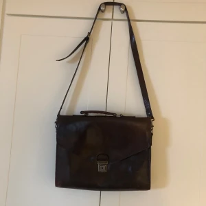 Brun skinnportfölj med rutigt foder - Säljer en stilren brun portfölj i skinn med ett klassiskt rutigt foder i brunt, gult och rött. Väskan har en justerbar axelrem och ett praktiskt handtag. Den är utrustad med en dragkedja på baksidan och ett lås framtill för säker förvaring. Br 38 H 28 Dj 6