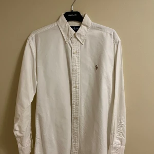 Vit skjorta från Ralph Lauren - Klassisk vit skjorta från Ralph Lauren i 100% bomull. Skjortan har långa ärmar och knappar framtill samt en broderad logotyp på bröstet. Perfekt för en stilren look.