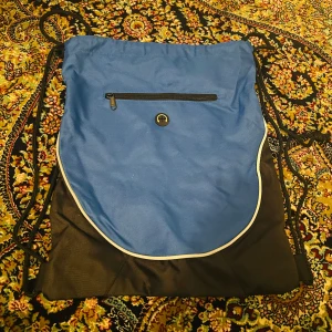 Blå och svart ryggsäck/ idrottspåse med dragkedja - Säljer en praktisk blå och svart ryggsäck/ idrottspåse med dragkedja framtill. Väskan har en nätpanel på baksidan och dragsko för enkel stängning. Perfekt för vardagliga äventyr eller träning.