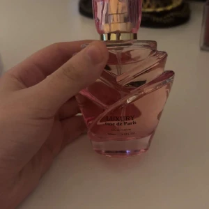 Luxury Rose de Paris Parfym - Elegant parfymflaska i rosa glas med en vriden design och gulddetaljer. Doften heter Rose de Paris och ger en lyxig känsla. Perfekt för den som älskar blommiga och sofistikerade dofter.