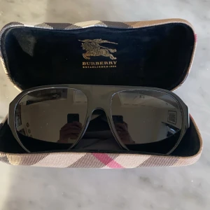 Svarta solglasögon från Burberry - Snygga svarta solglasögon från Burberry med en klassisk design. De har en solid svart båge och mörka linser, perfekt för soliga dagar. Kommer med ett stilrent fodral i Burberrys ikoniska rutmönster. Solglasögonen är i toppskick och sparsamt använda! Är det något ni undrar över är det bara att skriva!😊