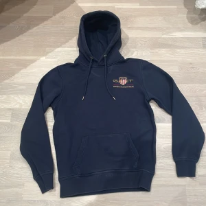 Mörkblå hoodie från GANT - Säljer en snygg mörkblå hoodie från GANT med broderad logga och text 'American Sportswear' på bröstet. Tröjan har en klassisk känguruficka och justerbar huva med snören. Perfekt för en avslappnad stil. 