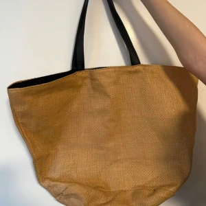 Beige shoppingväska med svarta handtag - Snygg och rymlig beige shoppingväska med svarta handtag. Väskan har en flätad design och är perfekt för sommarens äventyr. Insidan är fodrad och har en praktisk dragkedja för säker förvaring. Perfekt för att bära med sig allt man behöver under dagen.