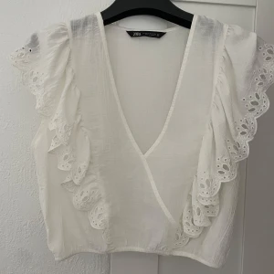Vit blus från Zara - Säljer en söt vit blus från Zara med volangdetaljer och broderade kanter. Blusen har en v-ringad design och korta ärmar med spetsdetaljer. Toppen är i nyskick, ALDRIG ANVÄND!☺️