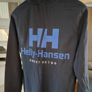 Långärmad tröja från Helly Hansen - Säljer en långärmad tröja från Helly Hansen med blå logotyp på bröstet och större tryck på ryggen. Perfekt för en avslappnad stil. Tröjan är i ett mjukt material och har en rund halsringning.