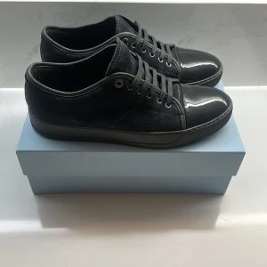 Svarta sneakers från Lanvin - Snygga svarta sneakers från Lanvin med en kombination av mocka och glansigt material på tån. Skorna har snörning och en stilren design som passar perfekt för en modern look. Pris kan diskuteras!
