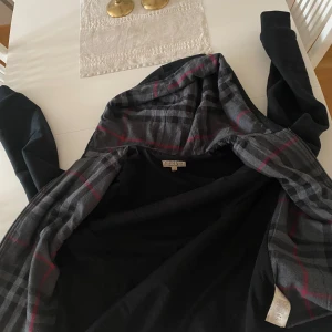 Svart hoodie från Burberry - Säljer en stilren svart hoodie från Burberry med en klassisk rutig insida i grått och rött. Den har en praktisk huva med snörning och långa ärmar. Perfekt för en avslappnad look. I storlek M. Nypris ligger på ca 4500