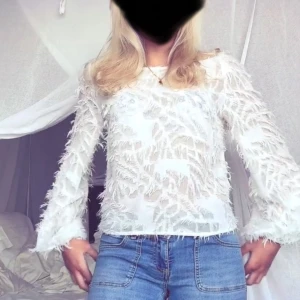 Vit blus med fjäderdetaljer - Säljer en vit blus från gina med fjäderliknande detaljer över hela tyget. Blusen har långa ärmar och en rund halsringning samt öppen rygg! jättefint skick